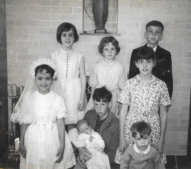 A rare capture of all eight Walsh kids in front of the fireplace on Claire’s First Communion day are in back (l-r) Teri, Bernadette and Thomas; in front are Claire, baby Francis held by Michael, and Denise and Joseph. WALSH FAMILY PHOTO